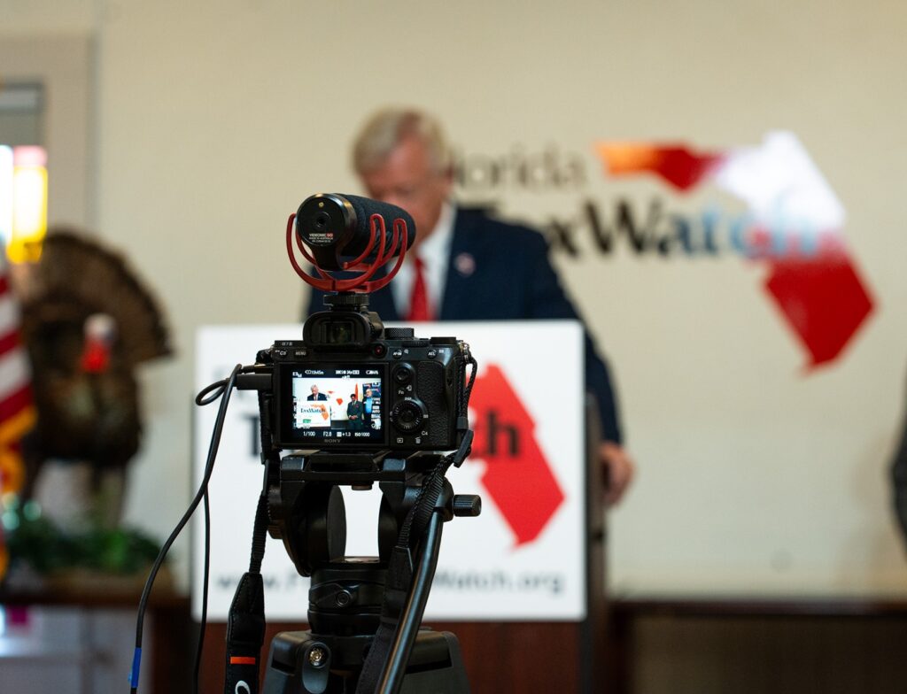 A Sony camera on a tripod with a red shotgun microphone films a speaker at a Florida TaxWatch press event. The camera's LCD screen shows the live shot in progress, with the Florida TaxWatch logo and branded signage visible in the background.